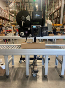 The SLAM system in use as a box passes down a roller conveyor and the machine applies a label to the package
