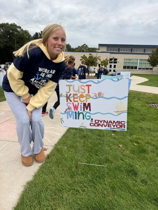 Amelia Coffin posing for photo next to Dynamic Conveyor sponsored sign at Walkathon event