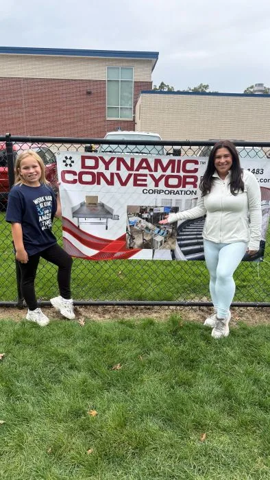 Lewis family mother and daughter posing by Dynamic Conveyor sponsor banner at Mona Shores School