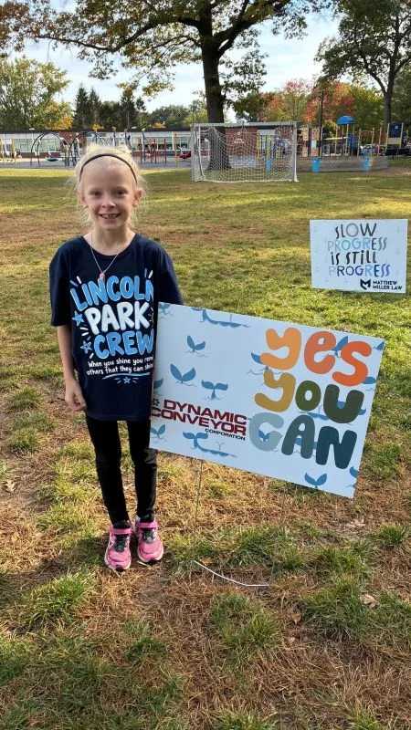 2026-walkathon-Student-Sign student standing by sponsor sign with Dynamic Conveyor logo